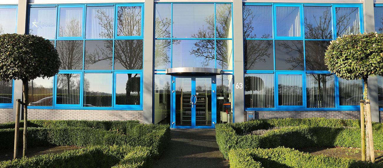 Moderne bedrijfsgevel met grote blauwe ramen, glazen entree en strak aangelegde groene tuin ervoor