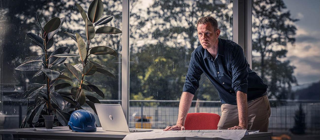 Persoon leunend over een bureau met laptop en bouwhelm in kantoorruimte met grote ramen op de achtergrond.
