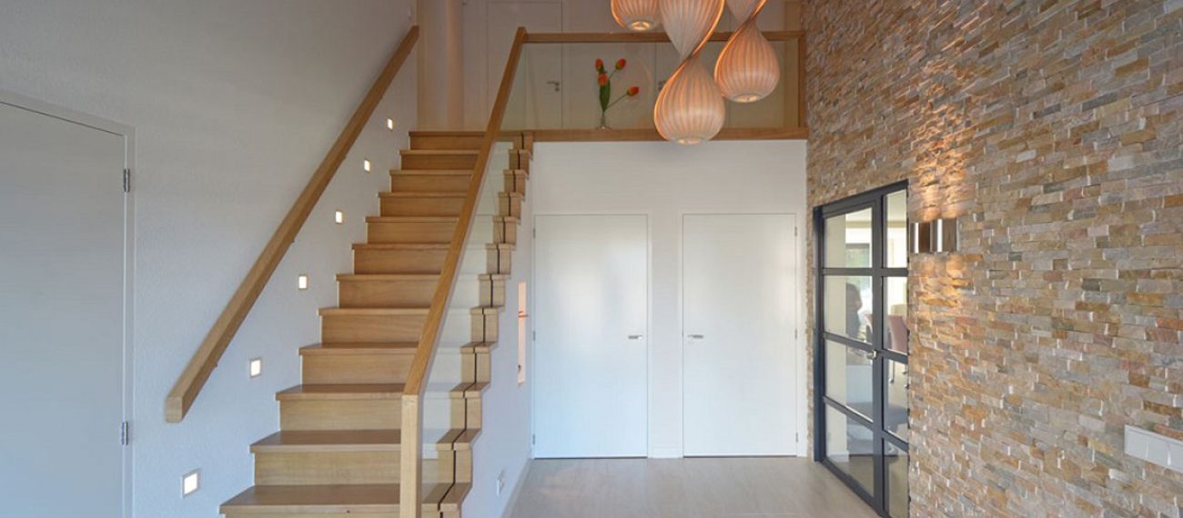 Entree van een moderne woning met houten trap, natuurstenen wand en glazen binnendeur.