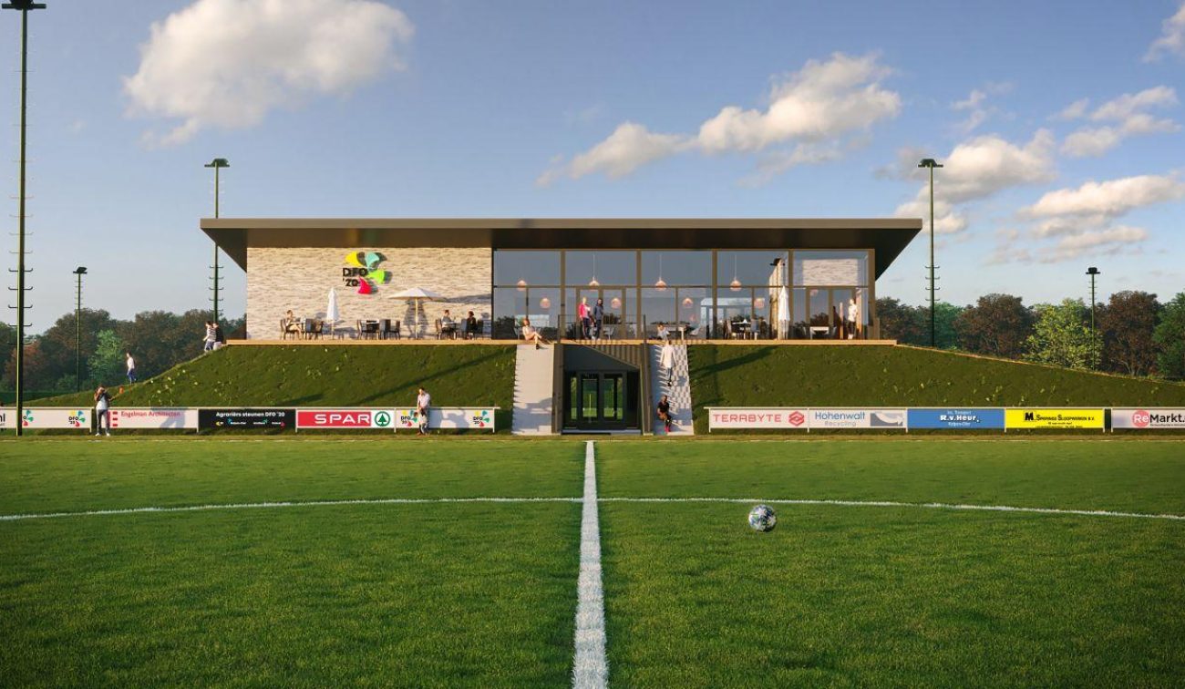 Artist impression van het nieuwe clubgebouw van DVO’20, frontaal gezien vanaf het hoofdveld, met glazen pui, terras en groene taluds; middenstip en reclameborden langs de lijn zichtbaar.