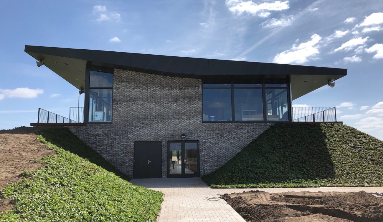 Voorzijde van het nieuwe clubgebouw DVO’20 met uitkragend dak, grote glaspuien en met klimop begroeide taluds; een klinkerpad leidt naar de entree in de bakstenen gevel onder een blauwe lucht.