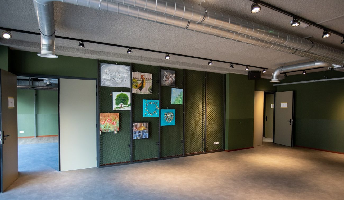 Interieur multifunctionele ruimte – groene wanden met gaaspanelen en kunstwerken, zichtbare luchtkanalen en spots aan plafond