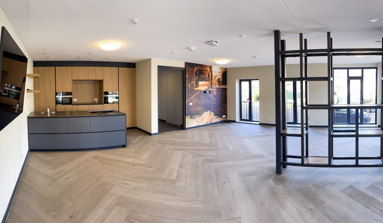 Interieur zorgcomplex Cicero Bleekstaete: ruime woonkamer met visgraatvloer, ingebouwde houtlook keuken, zwarte open vakkenkast als roomdivider, grote ramen en dubbele deuren naar balkon; wand met fotoprint en plafondspots.