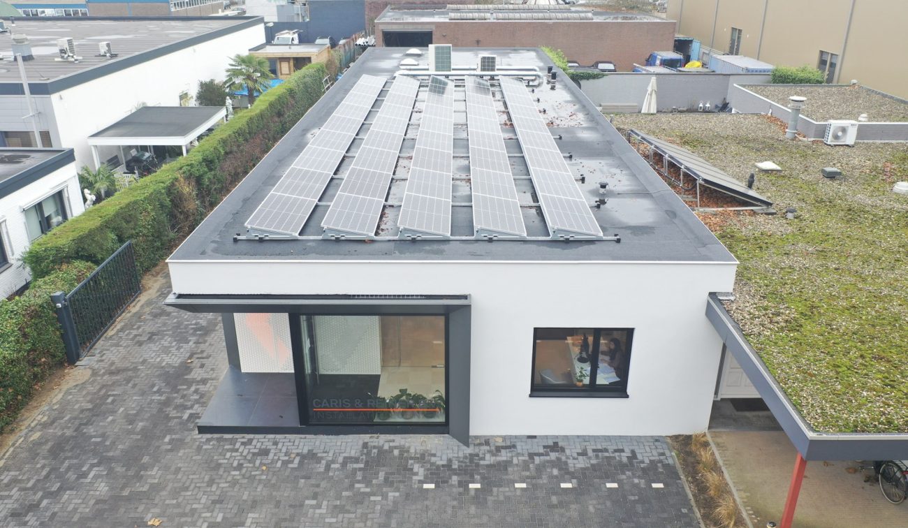 Luchtfoto van het gerenoveerde kantoor van Caris & Reiner met een plat dak vol zonnepanelen, witte gevel met zwart omlijste hoekentree en bestrate voorruimte; rechts een aangrenzend gebouw met groendak