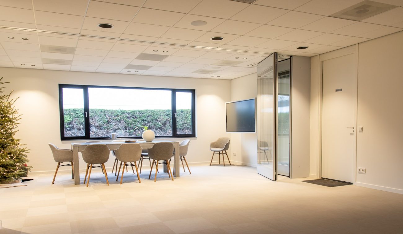 Lichte vergaderruimte met lange tafel en zes grijze stoelen bij een groot raam; rechts een wandmonitor, een opgevouwen glazen/akoestische scheidingswand en een witte deur.