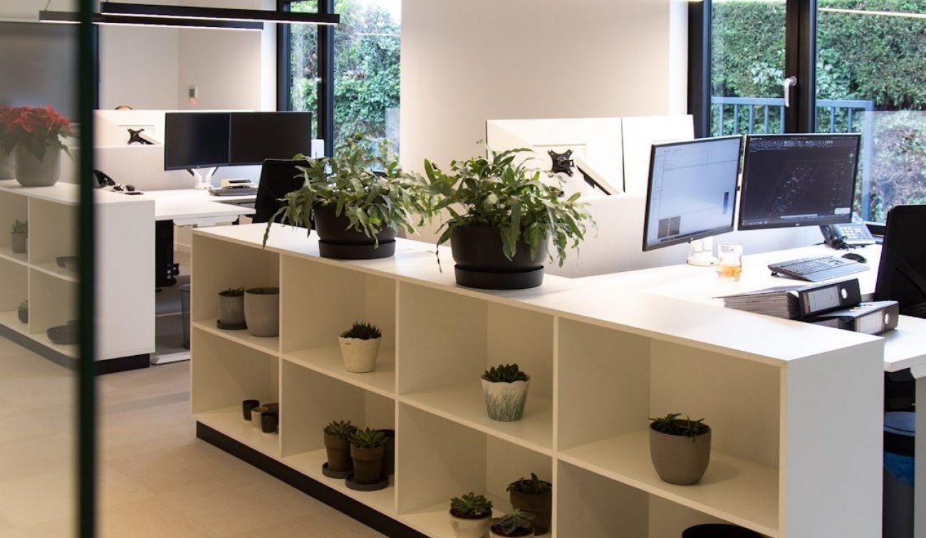 Lichte kantoorruimte met lange hanglampen, werkplekken met monitors en een witte open kast vol plantjes; door de ramen is een groene haag zichtbaar.