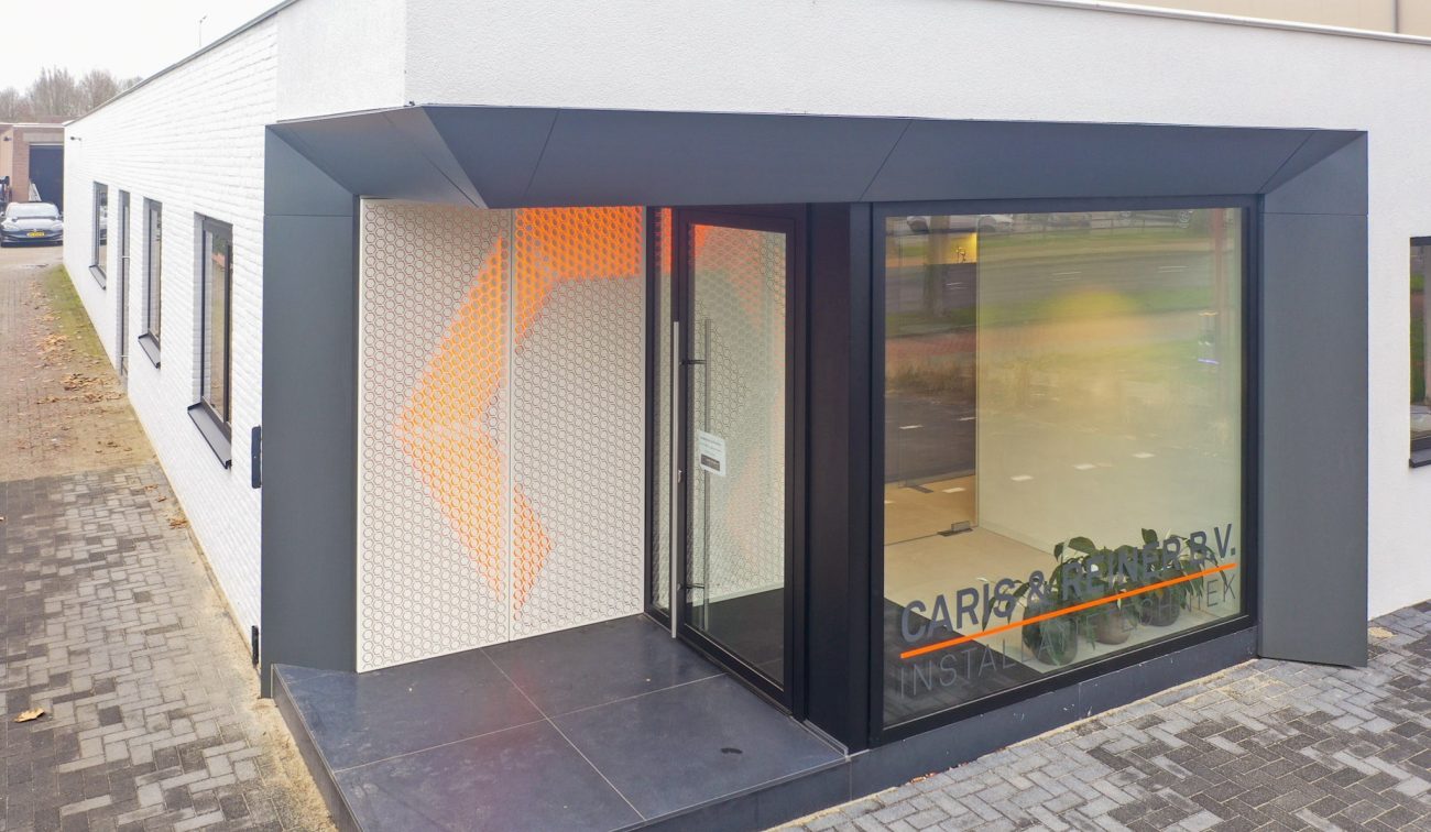 Hoekentree van gerenoveerd kantoor Caris & Reiner in Sittard met zwart kader, glazen pui en geperforeerde wand met oranje verlichting op een donkere tegelopstap.