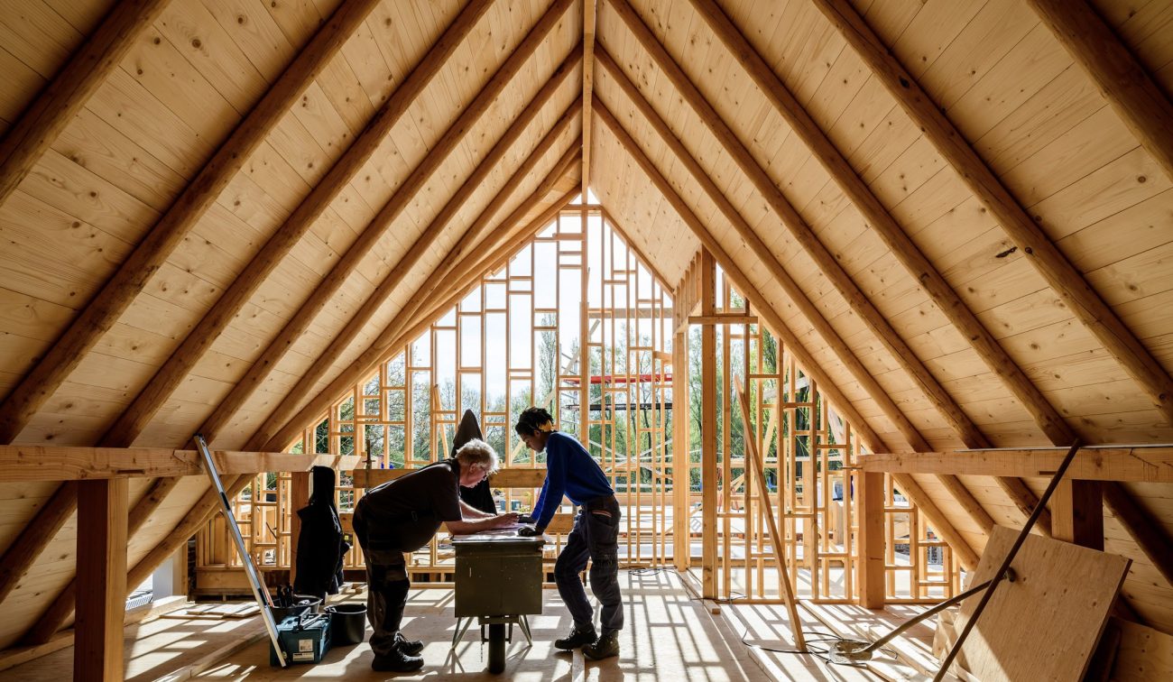 Zolder met houten spanten; twee vaklieden aan werkbank, houtskelet in aanbouw met veel daglicht