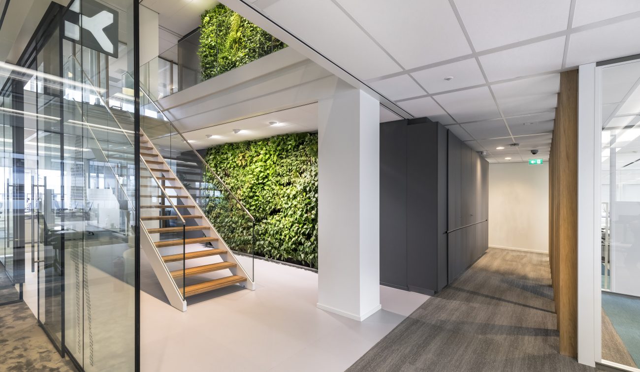 Renovatie kantoor: houten trap met glasbalustrade, groene plantwand en glazen vergaderruimtes, gang.