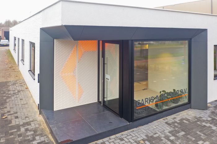 Hoekentree van gerenoveerd kantoor Caris & Reiner in Sittard met zwart kader, glazen pui en geperforeerde wand met oranje verlichting op een donkere tegelopstap.
