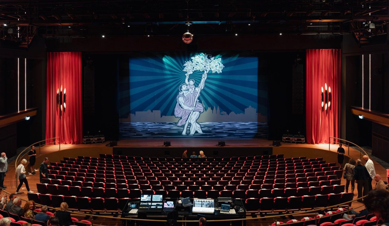 Theaterzaal De Oranjerie met rijen rode stoelen, een mengtafel achterin en een podium met rode zijgordijnen en projectie van een dansend paar met bloemen; bezoekers nemen plaats.