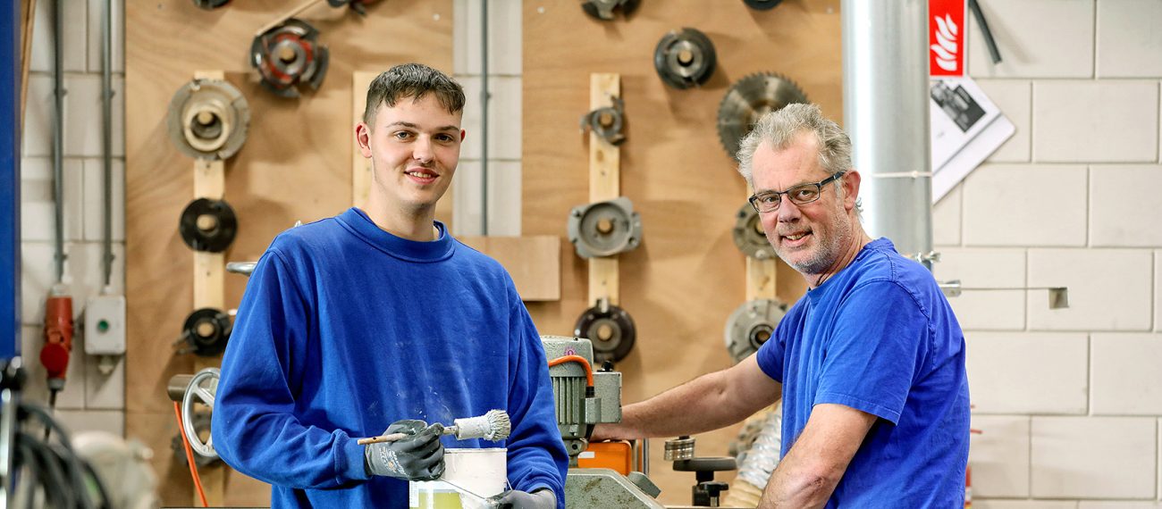 Twee medewerkers in een werkplaats staan bij een houtbewerkingsmachine; één houdt een verfbakje vast terwijl de ander aan de machine werkt, met gereedschap aan de muur op de achtergrond.