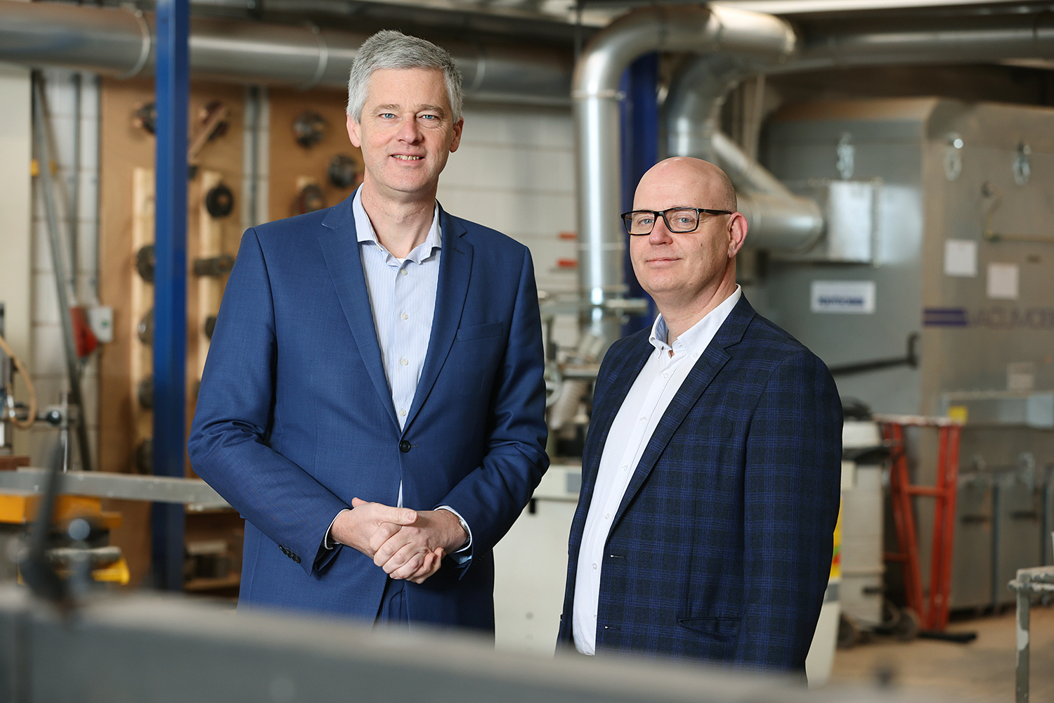 Twee mannen in zakelijke kleding staan samen in een technische werkplaats met installaties op achtergrond