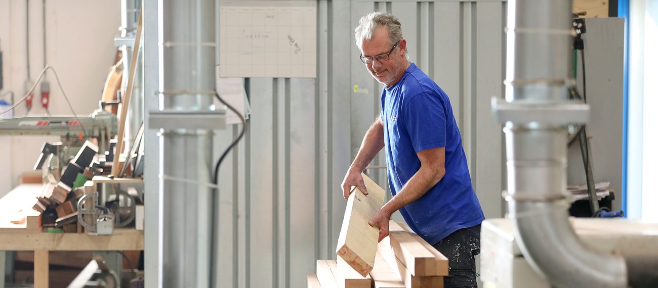 Een medewerker in een werkplaats stapelt houten balken, omringd door machines en gereedschappen.