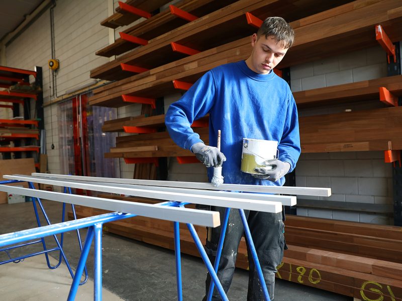 Persoon schildert lange houten latten in een werkplaats met rekken en hout op de achtergrond.