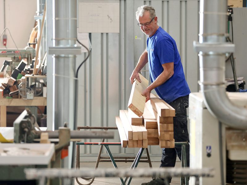 Een medewerker in een werkplaats stapelt houten balken, omringd door machines en gereedschappen.