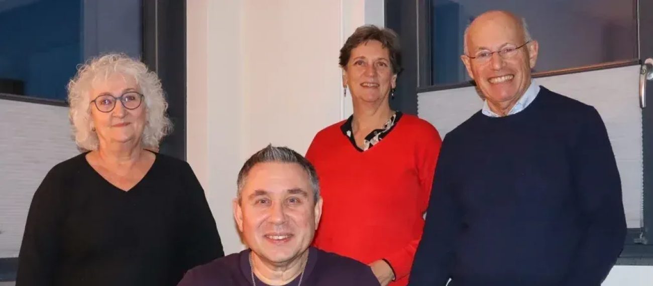 Vier volwassenen poseren binnen; man zit vooraan, drie staan achter hem en glimlachen naar de camera