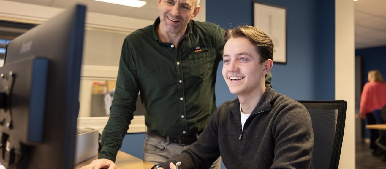 Twee collega’s kijken lachend naar een computerscherm terwijl één van hen aantekeningen maakt aan een bureau.