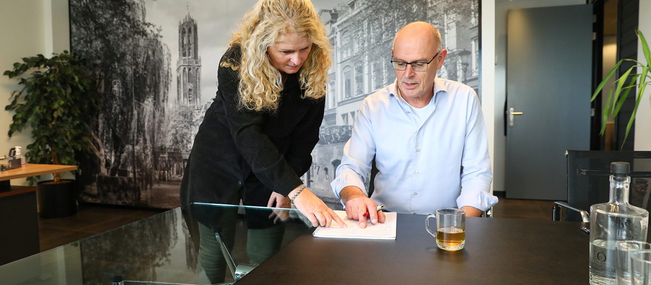 Twee collega’s overleggen boven notities aan vergadertafel met glazen blad en thee in modern kantoor