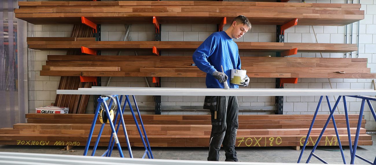 Een jonge man schildert lange houten latten in een werkplaats; hij houdt een verfbak in één hand en een kwast in de andere, met rekken vol hout op de achtergrond.