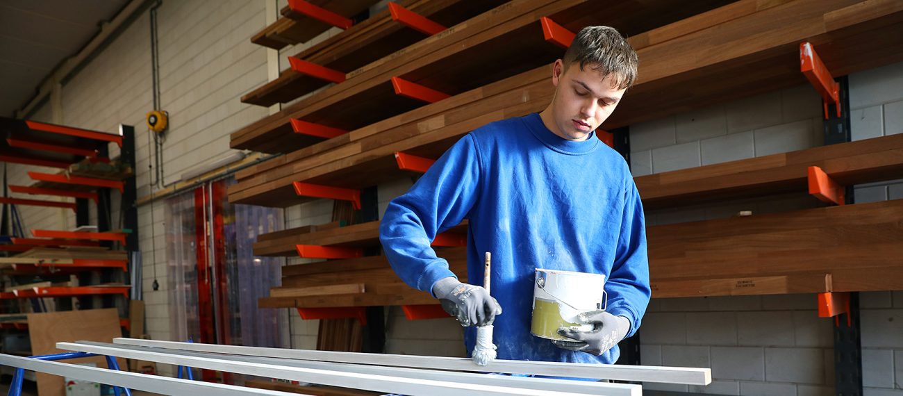Jonge medewerker in een werkplaats die houten latten verft met een kwast, met een verfblik in de andere hand en planken op de achtergrond.
