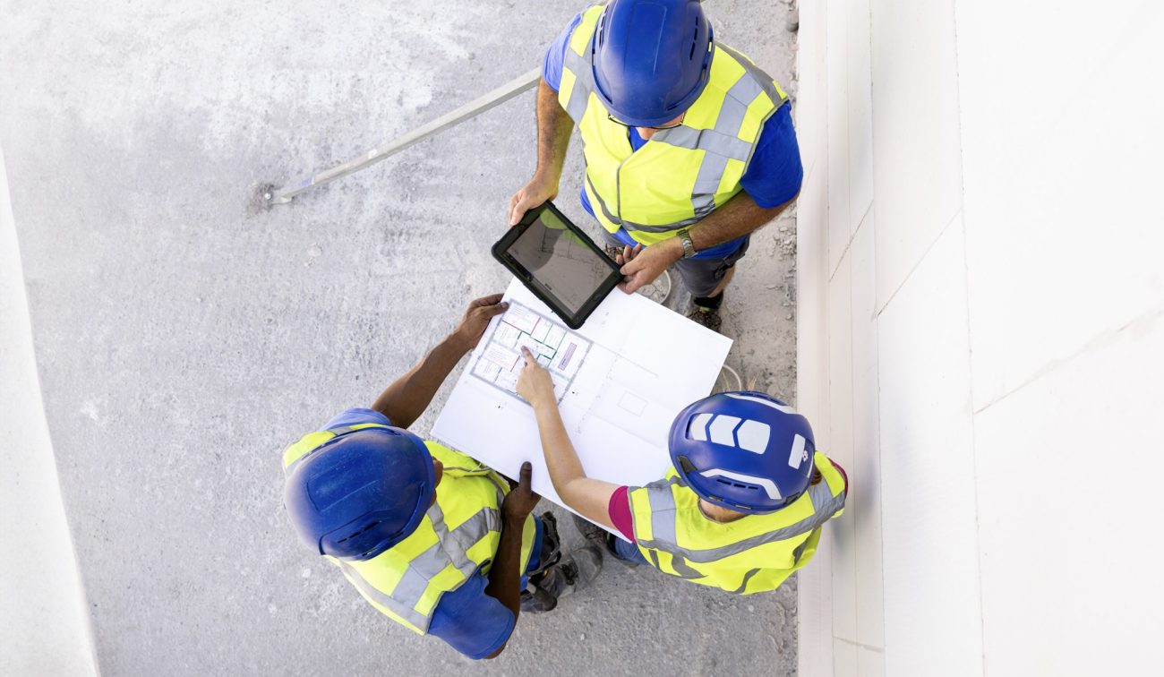 Drie bouwvakkers met veiligheidshelmen en reflecterende werkkleding bekijken documenten en tablet op bouwplaats