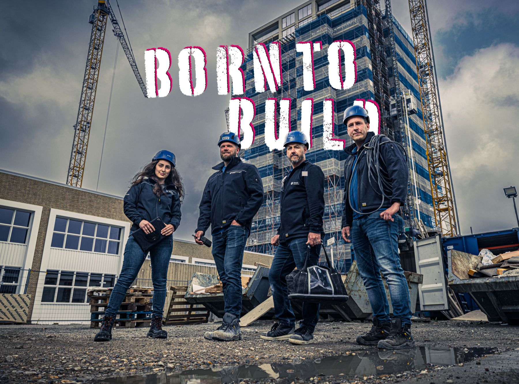Vier mensen met bouwhelmen poseren op een bouwplaats voor een hoge steigerconstructie, met de grote tekst ‘BORN TO BUILD’ boven hen in ruwe, opvallende letters.
