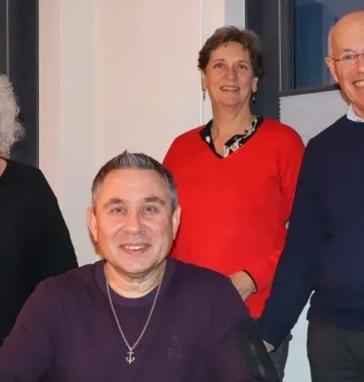 Vier volwassenen poseren binnen; man zit vooraan, drie staan achter hem en glimlachen naar de camera