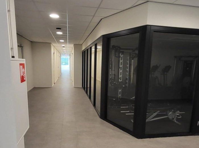 Transformatie voormalig hotel Born – gang met glazen wanden naar fitnessruimte, moderne verlichting en daglicht