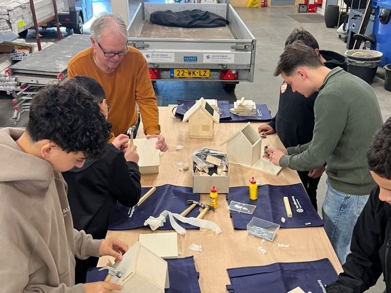 Studenten en begeleiders zijn aan een werktafel vogelhuisjes aan het timmeren