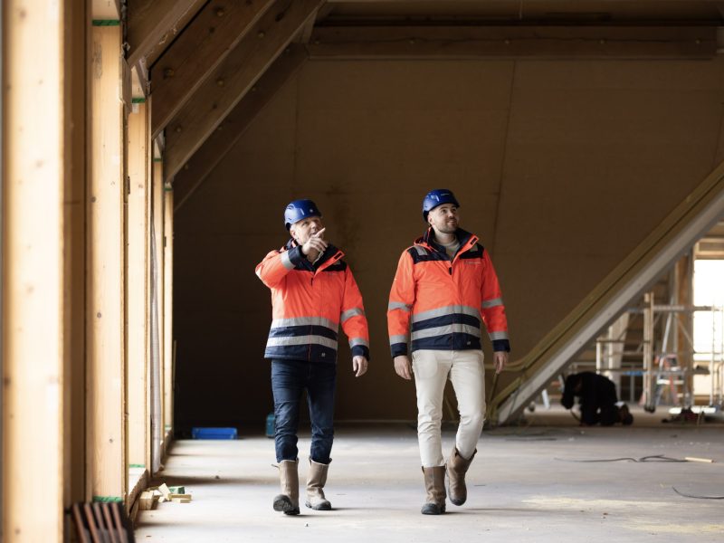 Twee collega's met bouwhelmen en veiligheidsjassen aan lopen door een gebouw in aanbouw, de linker persoon wijst iets aan