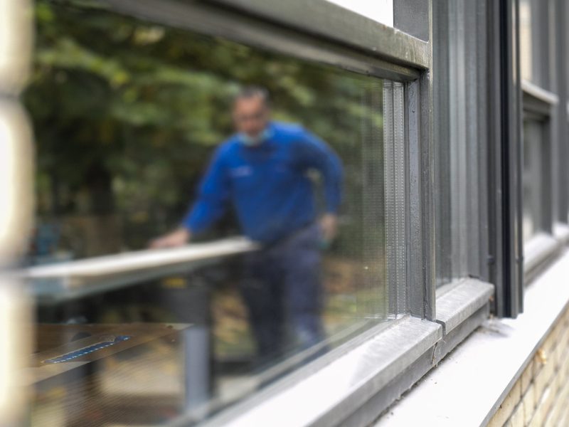 Close-up van grijs aluminium kozijn met dubbele beglazing; buiten in de onscherpe achtergrond werkt een monteur in blauwe trui aan een zaagtafel.