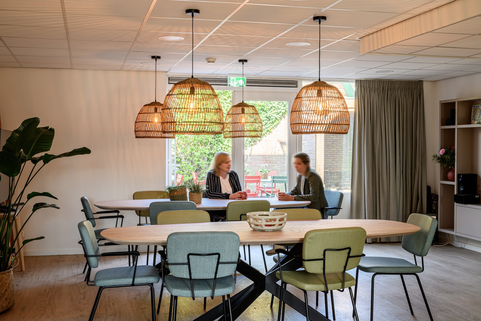 Gemeenschappelijke ruimte met houten tafels, groene stoelen, hanglampen en uitzicht op binnentuin