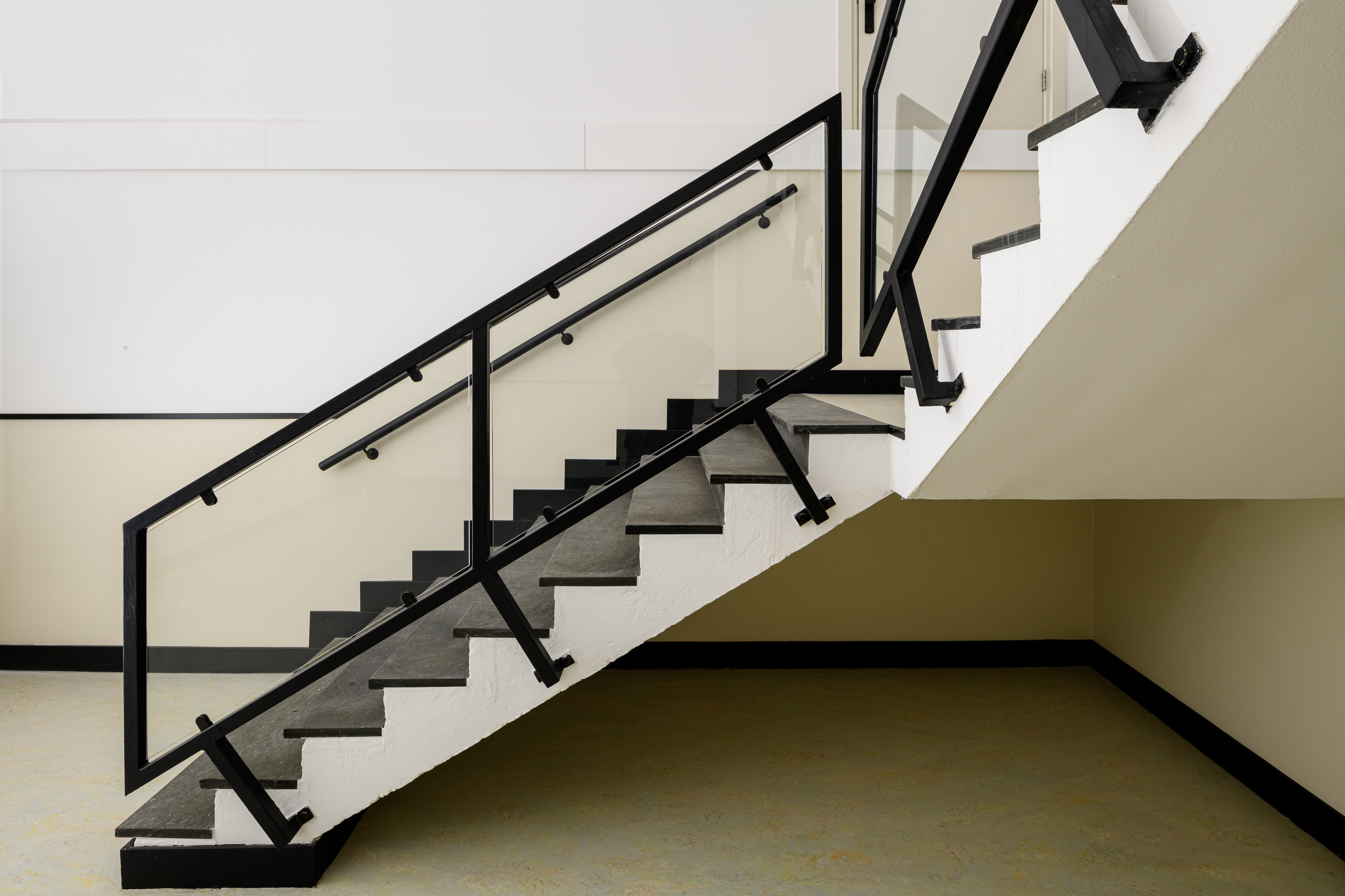 Moderne schooltrap: glaspanelen, dubbele handregel langs de muur en grijze steen-/rubbertreden.