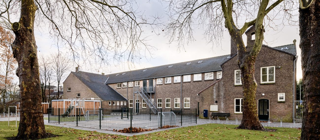 Gerestaureerd schoolgebouw in baksteen met speelplaats, hekwerk en twee grote platanen op voorgrond.