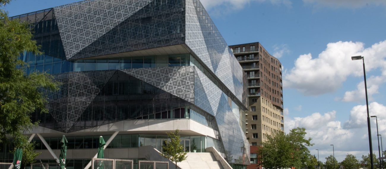Hoekig glazen volume boven transparante plint; plein met plantenbakken en toren op achtergrond.