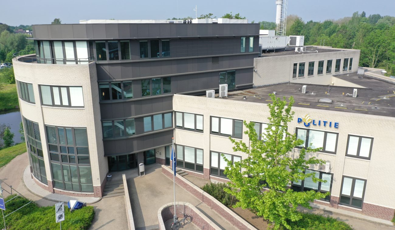 Luchtfoto van het gerenoveerde politiebureau in Roermond met ronde hoekgevel, donkergrijze bovenbouw, plat dak met installaties, ‘Politie’-logo op de gevel, vlaggenmast en boom in de voorgrond.