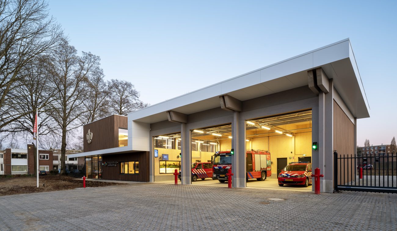 Nieuwe brandweerpost Sittard-Oost bij schemering met drie open uitrijpoorten; binnen staan een brandweerwagen en dienstauto klaar, naast de glazen entree in het moderne hout-wit gebouw.