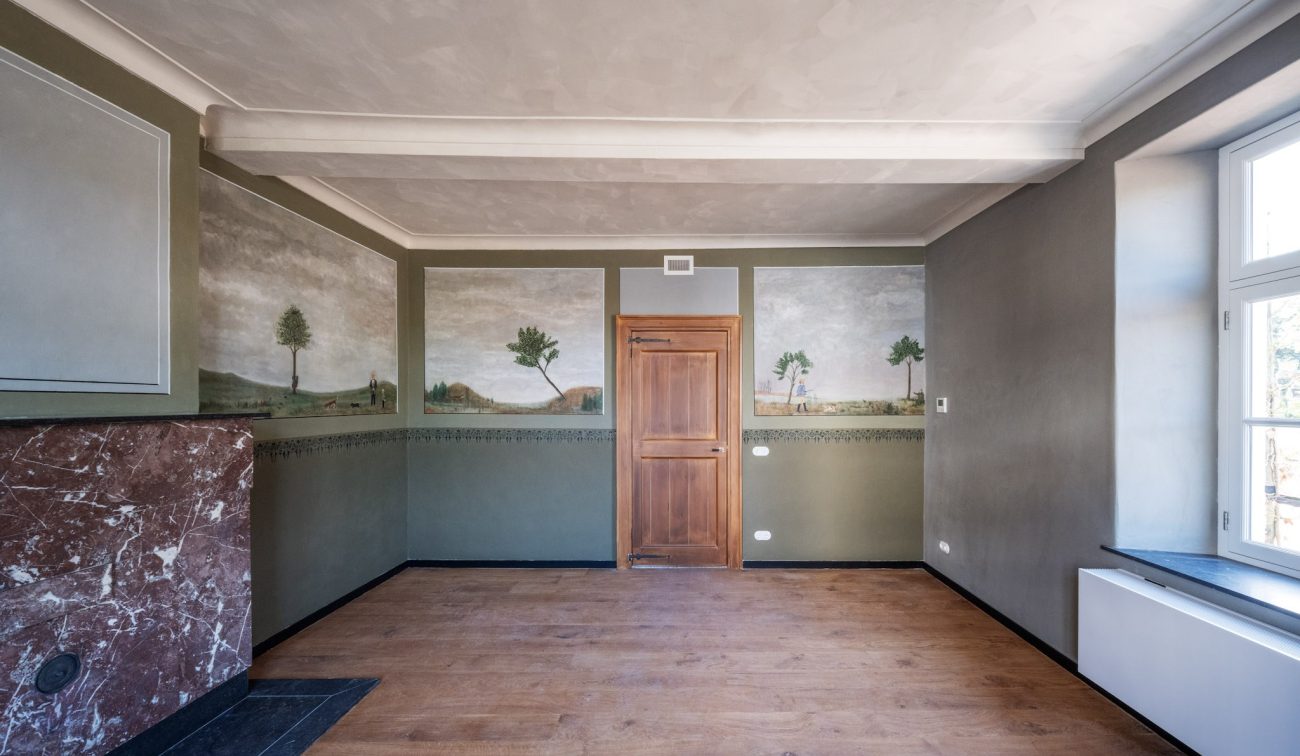 Gerestaureerde kamer in de Grathemermolen met houten vloer, groen-grijze wanden met landschappelijke muurschilderingen, houten deur centraal, marmeren schouw links en groot raam rechts.
