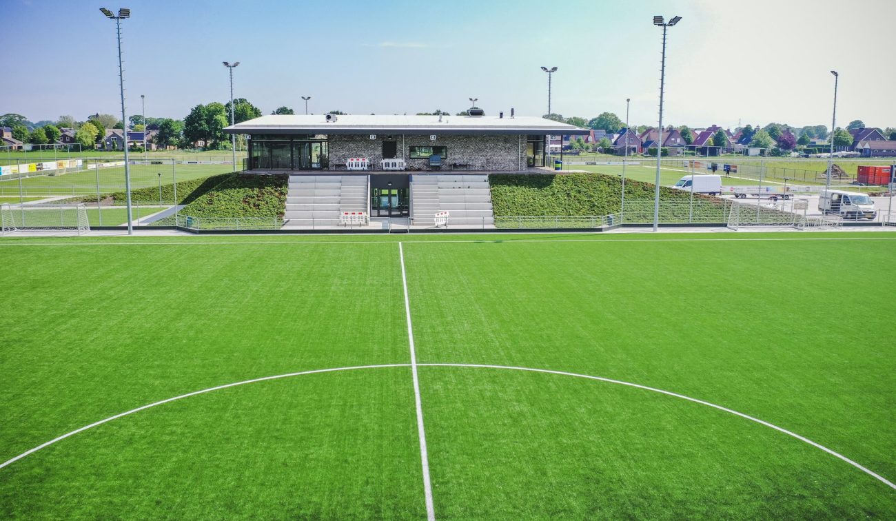 Nieuw clubgebouw DVO’20 met geïntegreerde tribune en sedumdaken, gezien vanaf het kunstgrasveld met middenstip.