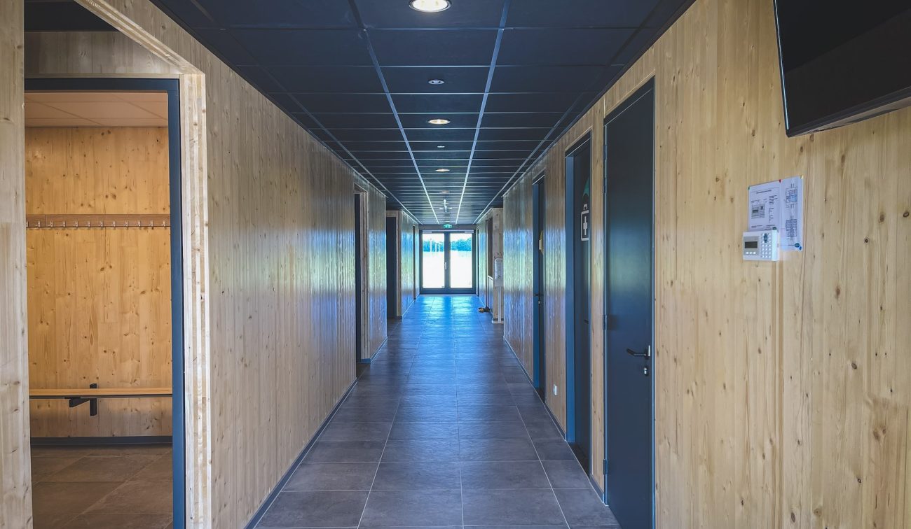 Gang in clubgebouw DVO’20 met houten wanden, donker plafondsysteem en grijze tegelvloer, met deuren naar kleedkamers en lichtinval bij de uitgang.