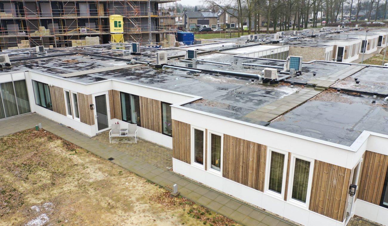 Nieuwbouw zorgwoningen Amstenrade – laagbouw met witte gevels en houten lamellen, plat daken vol installaties, betegeld pad met bankje; op de achtergrond bouwsteiger en aangrenzende paviljoens.