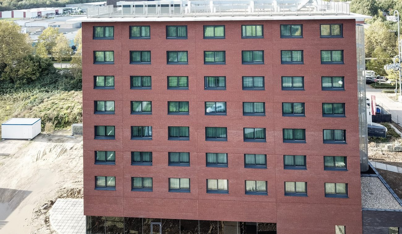 Voorgevel van het nieuwe Bastion Hotel in Geleen: rood­bakstenen blok met een strak raster van donkere, rechthoekige ramen over acht verdiepingen; op het platte dak staan installaties. Aan de voet van het gebouw is nog bouwterrein zichtbaar.