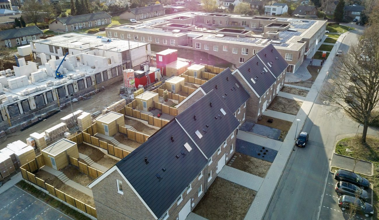 Luchtfoto nieuwbouw sociale eengezinswoningen in Hulsberg met rijwoningen met zwarte zadeldaken, achtertuinen en naastgelegen bouwplaats met appartementen in aanbouw.