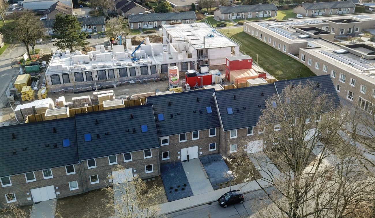 Luchtfoto nieuwbouw sociale eengezinswoningen in Hulsberg: rij bakstenen huizen met zwarte zadeldaken langs de straat, met achtertuinen; daarachter een bouwplaats met witte wanden en rode bouwcontainers.