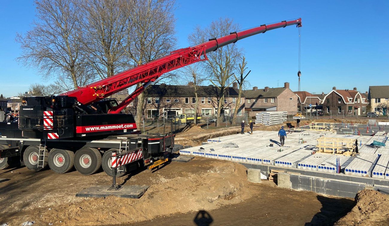 Bouwplaats nieuwbouwwoningen – mobiele telescoopkraan plaatst kanaalplaatvloeren op fundering, monteurs aan het werk bij blauwe lucht