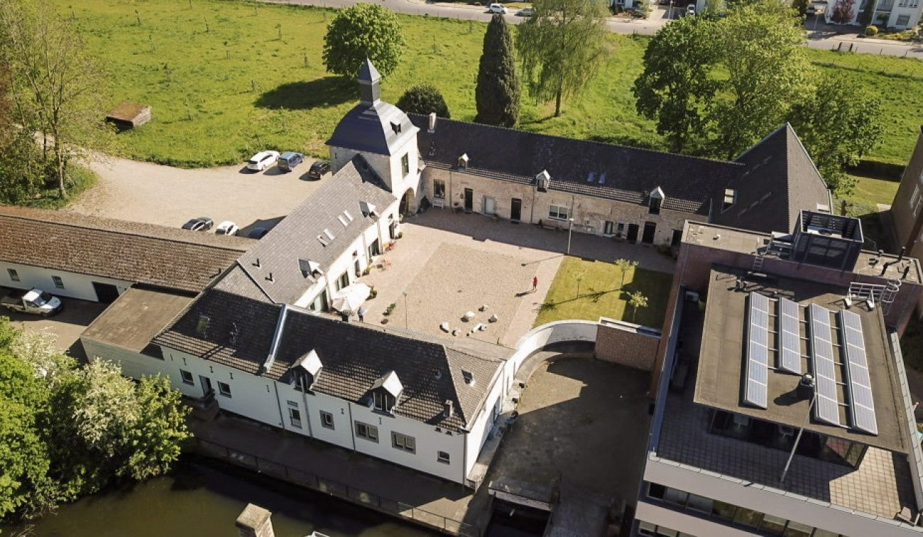 Luchtfoto carréhoeve met torentje en grindbinnenplaats aan het water, naast modern gebouw met zonnepanelen — Herbestemming hoeve tot appartementen Maastricht