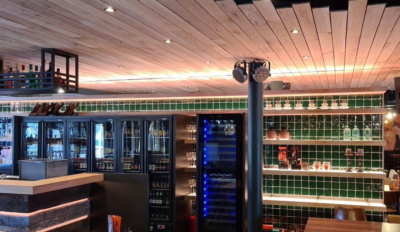 Interieur eetcafé De Aanwinst: barwand met koelvitrines en open schappen met groen tegelwerk, houten plafond en staande spot.