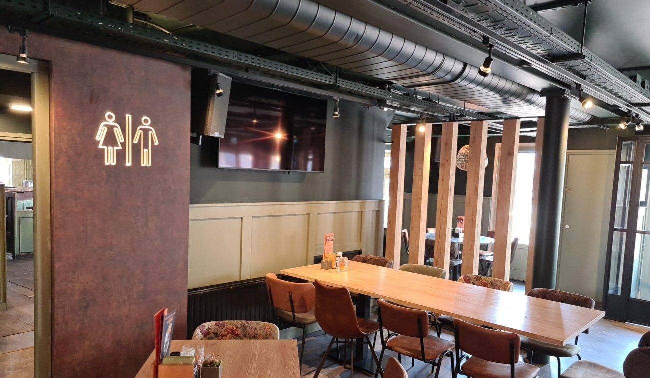 Gezellige eetcafé-ruimte met lange houten tafel, leren stoelen en industrieel plafond met zichtbare luchtkanalen.