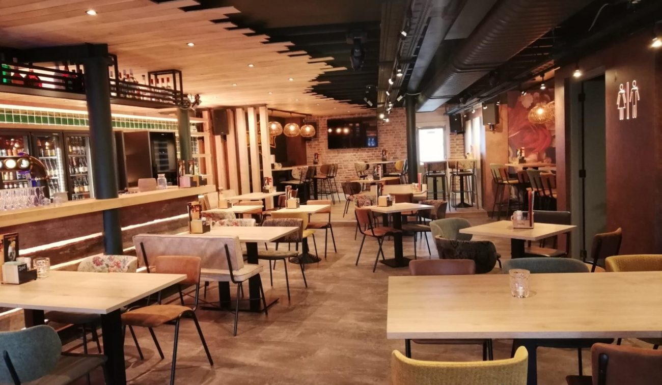 Interieur van eetcafé De Aanwinst in Nederweert: warme zithoek met houten tafels en diverse stoelen, lange bar met flessenkoelingen links, industriële zwarte luchtkanalen aan het plafond en toiletpictogram aan de rechterwand.
